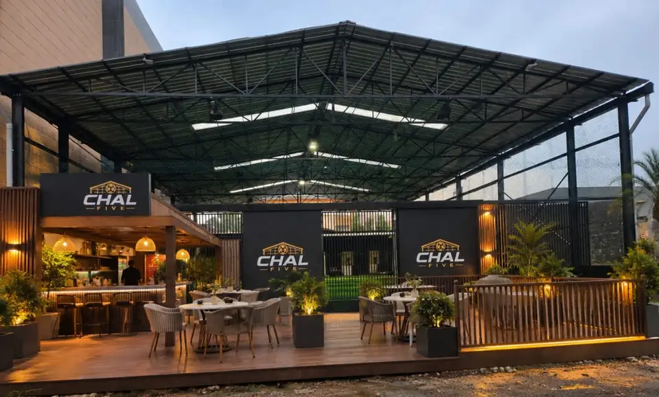 La terrasse de CHAL Five en début de soirée&nbsp;: tables, fauteuils, plantes éclairées et bar, devant le terrain couvert.